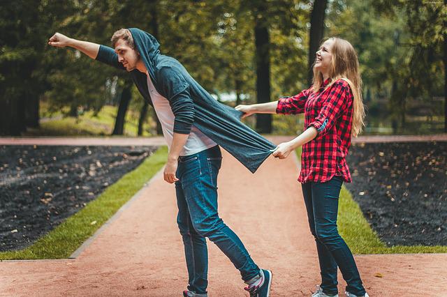 Two people in a park one in front of the other. The person behind is pulling the jacket of the person in front who is doing a superhero pose