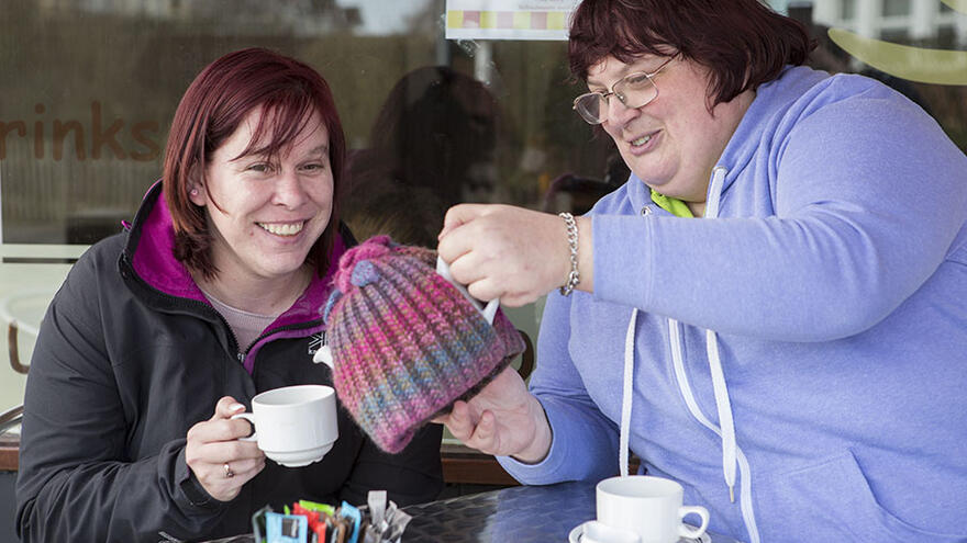 Two women sat at a table outside a cafe. One woman is pouring the other a cup of tea from a teapot.