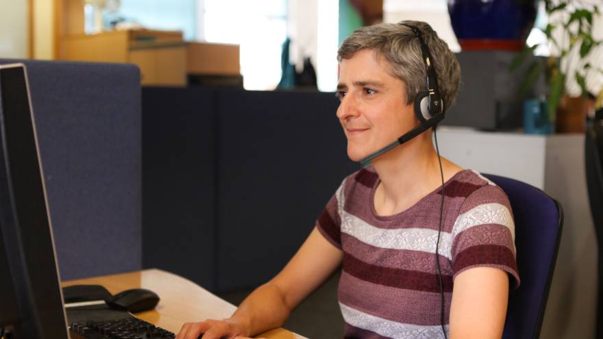 Woman with short grey and purple striped top. She is sat at a desk in an office with a computer on it. She is smiling whilst wearing a telephone headset.
