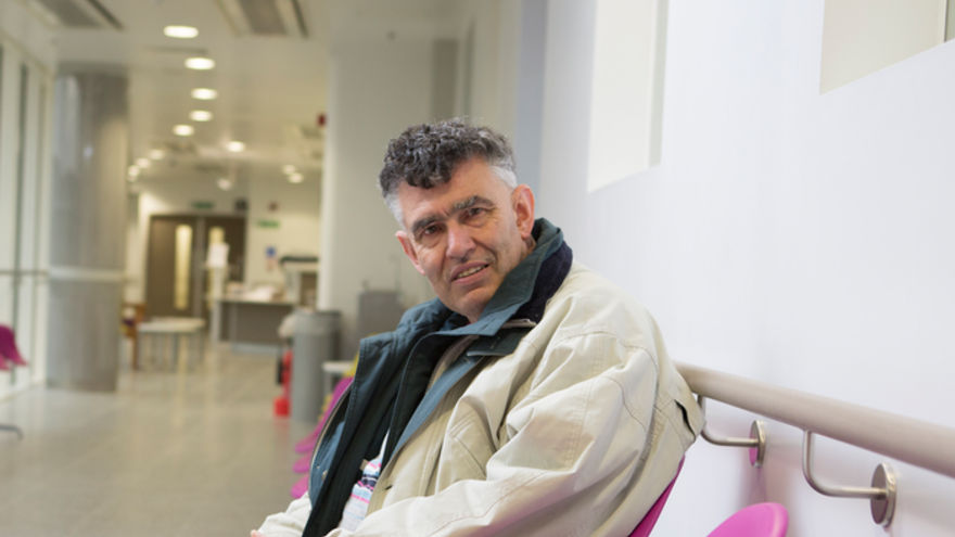 Older gentleman wearing overcoat sat smiling in hospital corridor.