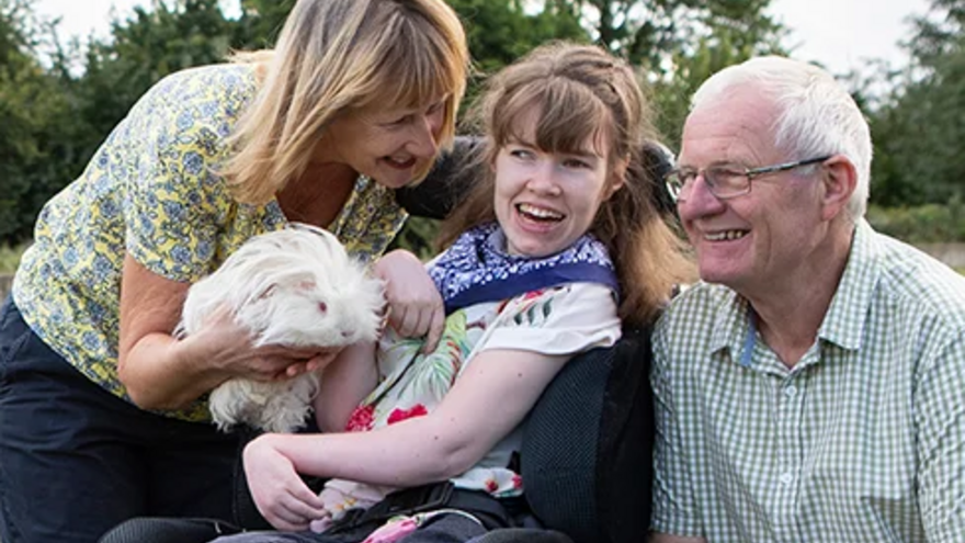 Mother and father stood with their daughter who is using a wheelchair. They are in a garden next to an animal pen, the mother is holding a guinea pig for her daughter to look at.