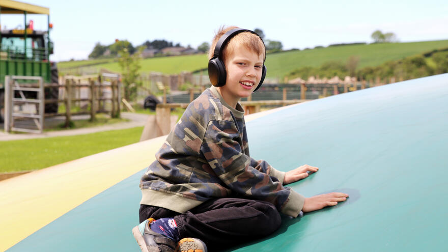 A boy wearing ear defenders kneels down onto a trampoline at an accessible playground.