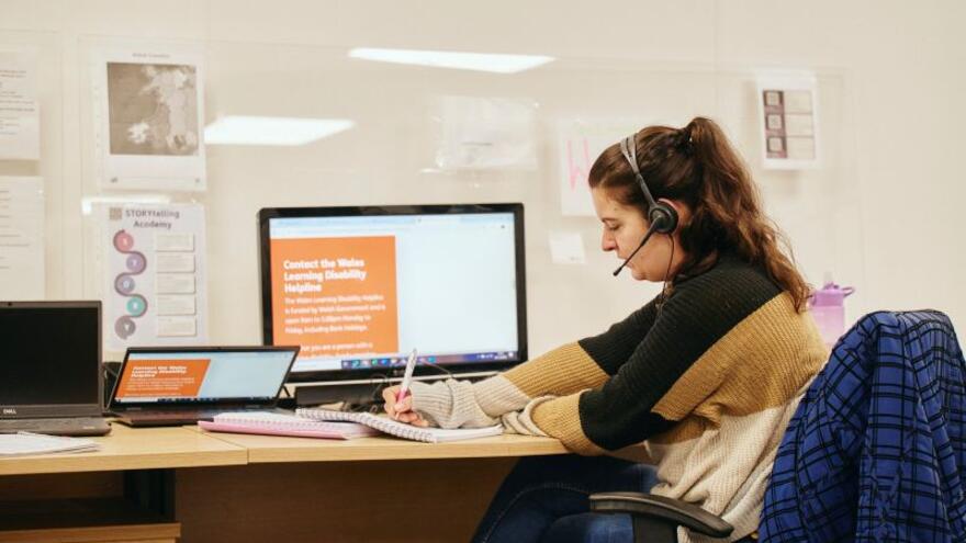 Helpline caseworker answering a helpline question while sat at a desk. The woman is wearing a headset, is sat at a desk with a computer and is writing notes