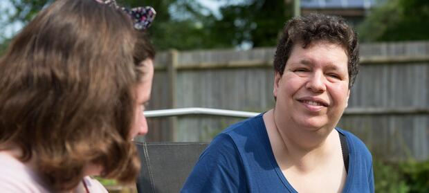 Woman wearing dark blue t-shirt, with dark hair, sat outside in front of garden fence, smiling at camera