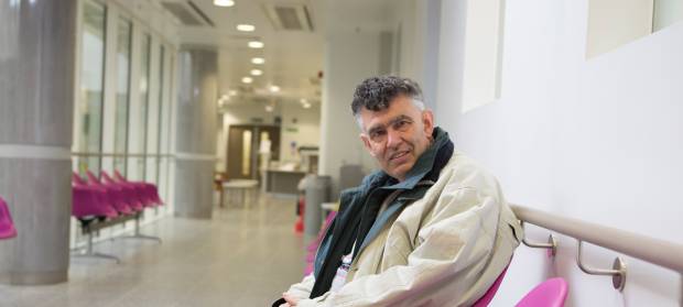 Older gentleman wearing overcoat sat smiling in hospital corridor.