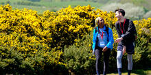 Man and woman walking together next to yellow-flowering bushes.
